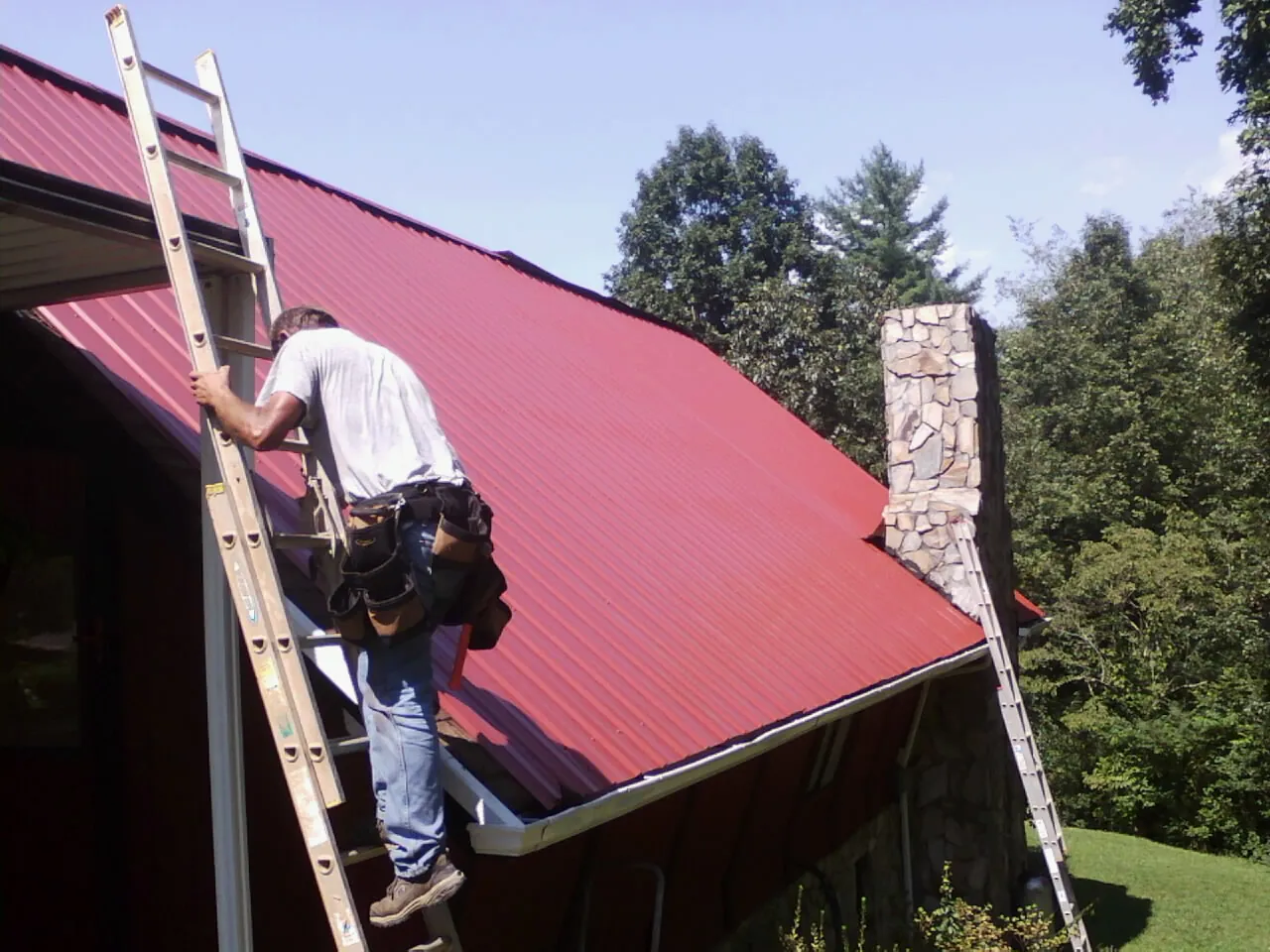Roofer on a ladder beside a red metal roof and stone chimney — Soffit & Fascia Repair by Roofing Brothers in Schooleys Mountain